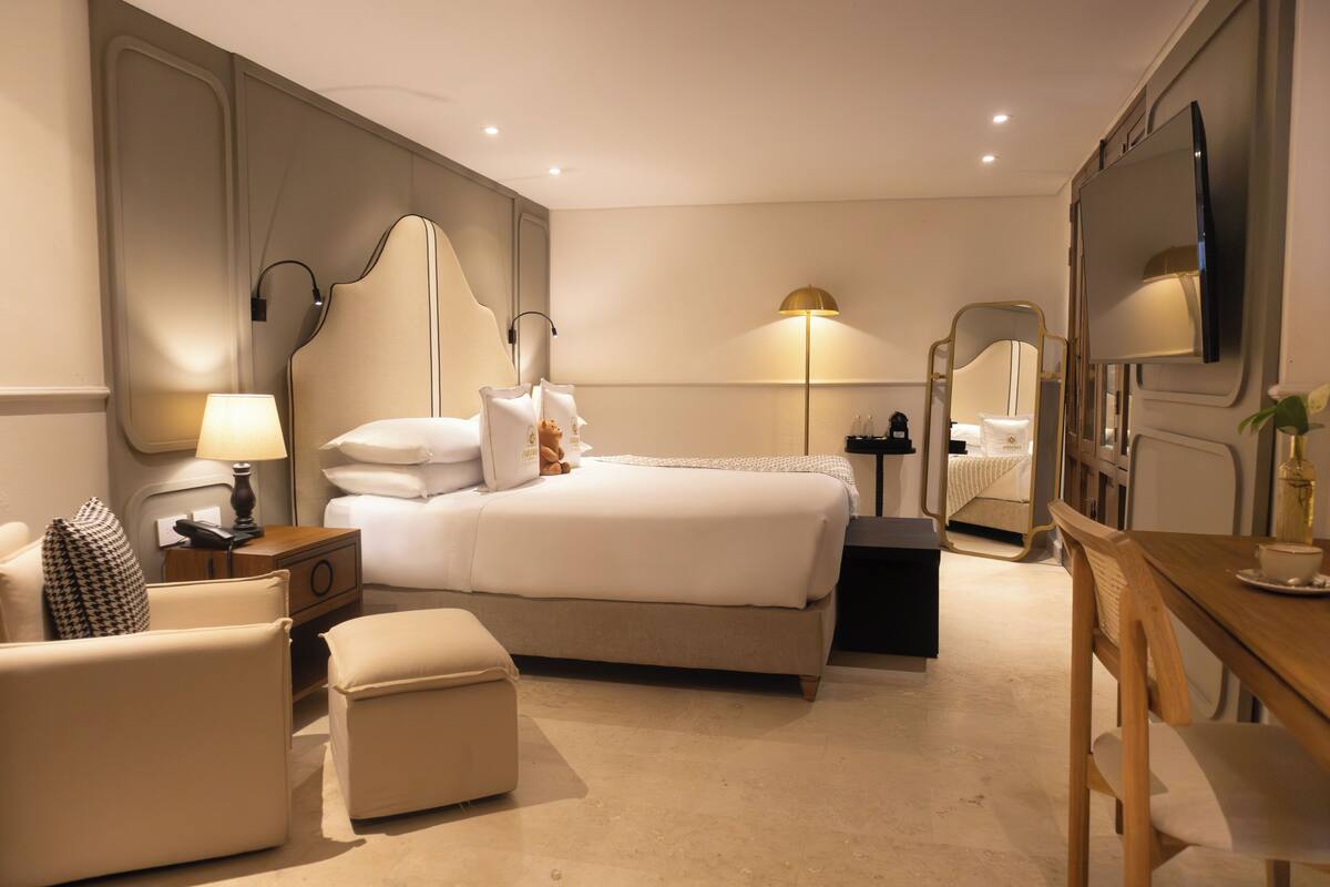 Habitación de hotel con cama, mesita de noche, lámpara, sillón, puf y escritorio con silla.
