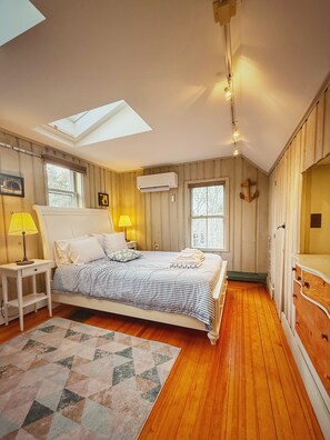 A bedroom with a skylight, a bed with a striped bedspread, a bedside lamp, a wooden floor, and a window with a view of trees. - Chatham Family Retreat w/ Heated Pool (Chatham)