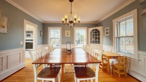 A dining room with a large wooden table, white chairs, and a chandelier. There are windows on the right side, and a kitchen is visible on the left. - Chatham Family Retreat w/ Heated Pool (Chatham)