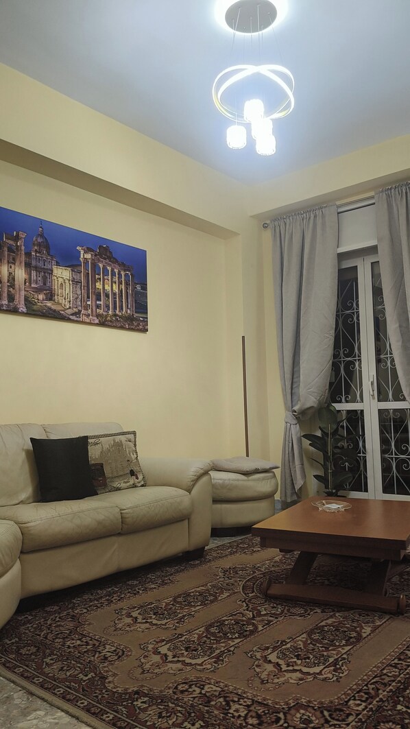 Een woonkamer met een beige bank, een salontafel, een kamerkleed met patroon, een schilderij van een historisch gebouw en een raam met gordijnen.