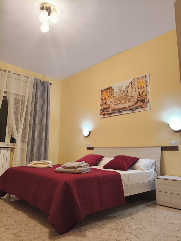 Habitación de hotel con cama, mesita de noche, lámpara de pared, un cuadro y una ventana con cortinas.