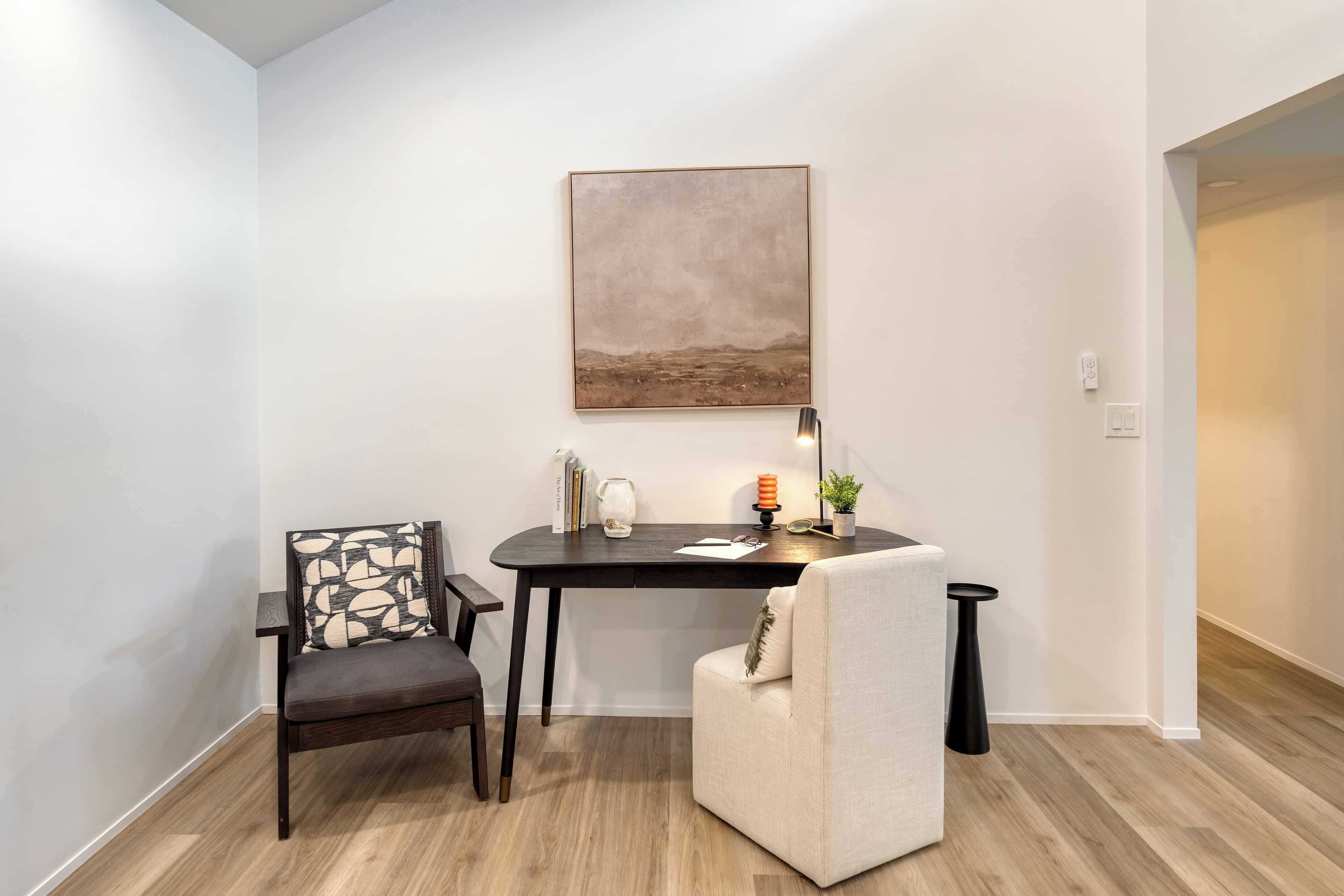 Une pièce moderne avec un bureau en bois foncé, une chaise dotée d’un coussin à motifs, une petite table ronde avec une lampe et un tableau accroché au mur.
