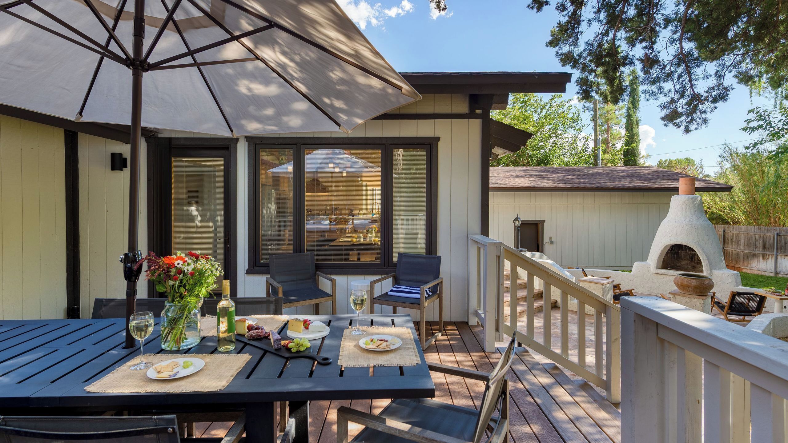 An outdoor patio with a table set for dining, a chair, a large umbrella, and a wood-burning oven.