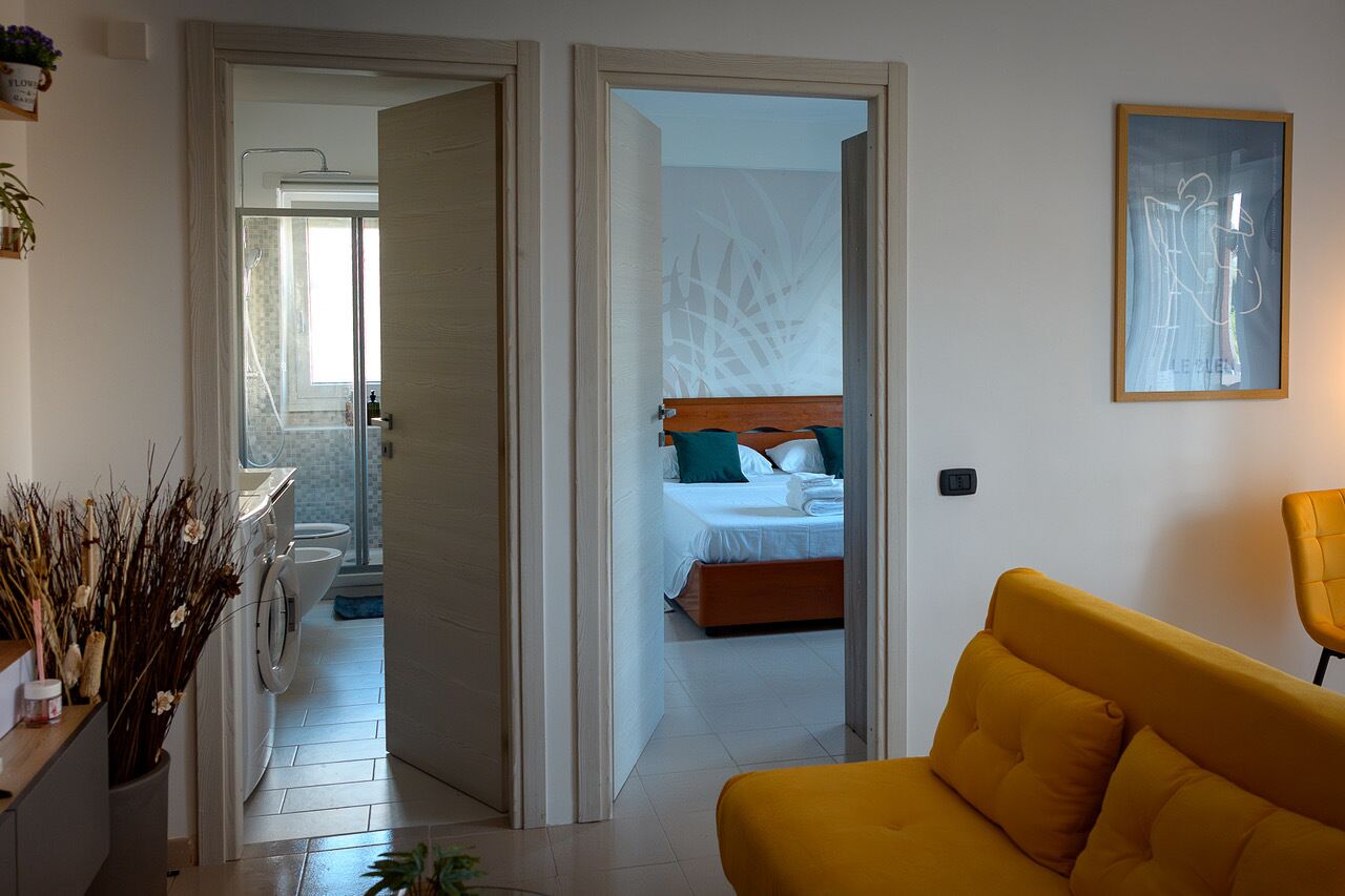 A hotel room with a bed, a yellow sofa, a bathroom with a shower, and a hallway leading to another room.