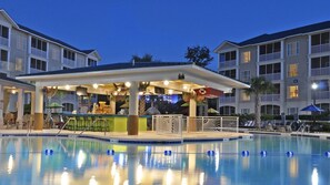 Eine Poolbar mit überdachtem Sitzbereich, ein Schwimmbecken mit Bahnen und angrenzende mehrstöckige Gebäude.