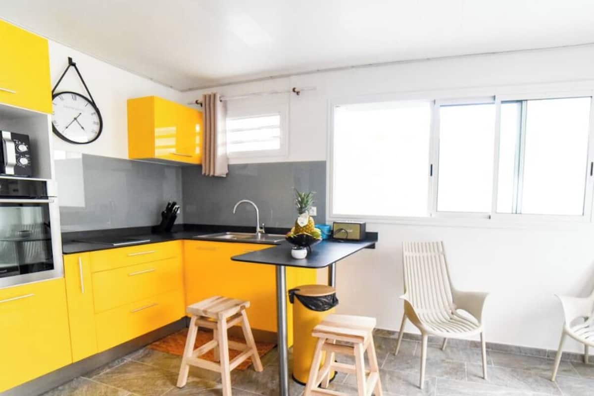 Una cocina moderna con armarios amarillos, encimera negra y un pequeño comedor con dos taburetes y una silla.