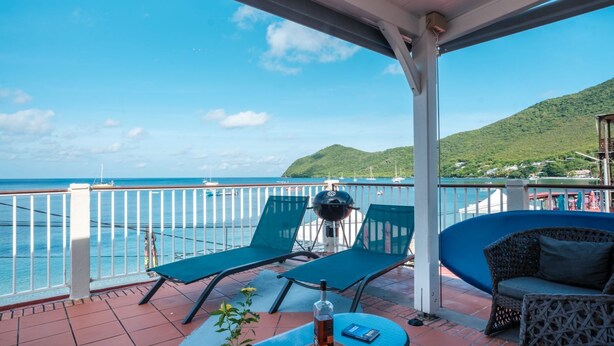 A balcony with two lounge chairs, a small table, and a grill. The balcony overlooks a body of water with boats and a hilly landscape in the background. - LEZ'ARLET PLAGE  (Les Anses-d'Arlets)