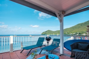 A balcony with two lounge chairs, a small table, and a grill. The balcony overlooks a body of water with boats and a hilly landscape in the background. - LEZ'ARLET PLAGE  (Les Anses-d'Arlets)
