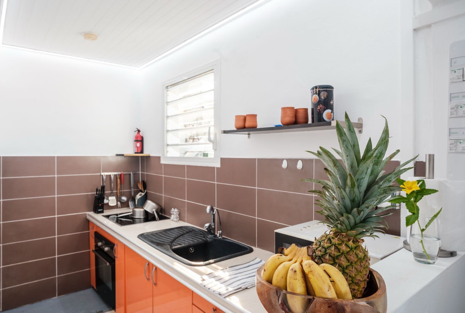 A kitchen with brown and white tiles, a sink, a stove, and a window. There are kitchen utensils and a fire extinguisher on the counter.