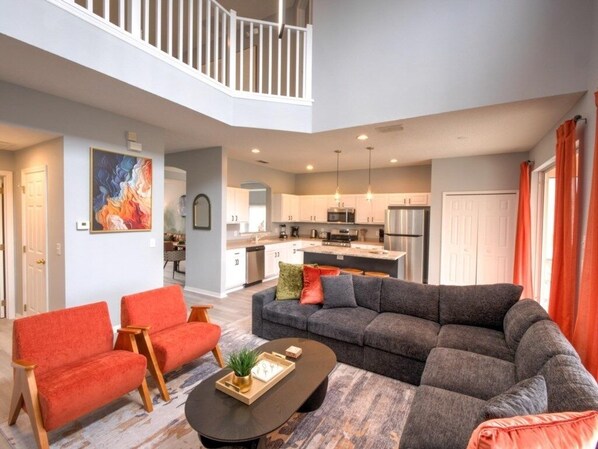 A spacious living room with a gray sectional sofa, two red armchairs, and a coffee table. A kitchen is visible in the background with modern appliances.
