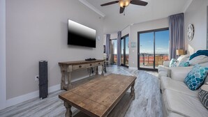 A modern living room with a flat-screen TV, a wooden coffee table, a sofa with patterned cushions, and a balcony with a view. - Phoenix X 102 1 Bedroom Condo (Orange Beach)