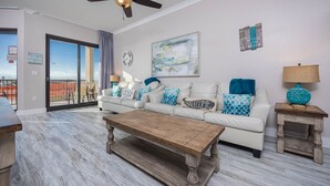 A living room with a white sofa, a wooden coffee table, and a view of the ocean through a sliding glass door. - Phoenix X 102 1 Bedroom Condo (Orange Beach)