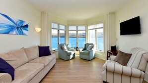 A living room with a beige sofa, two armchairs, a coffee table, and a TV. Large windows offer a view of the water. - Dean Lake House (Watkins Glen)