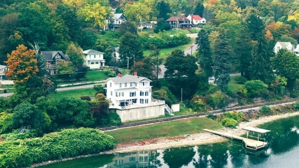 An aerial view of a large white house by a lake, surrounded by lush greenery and other houses. - Dean Lake House (Watkins Glen)