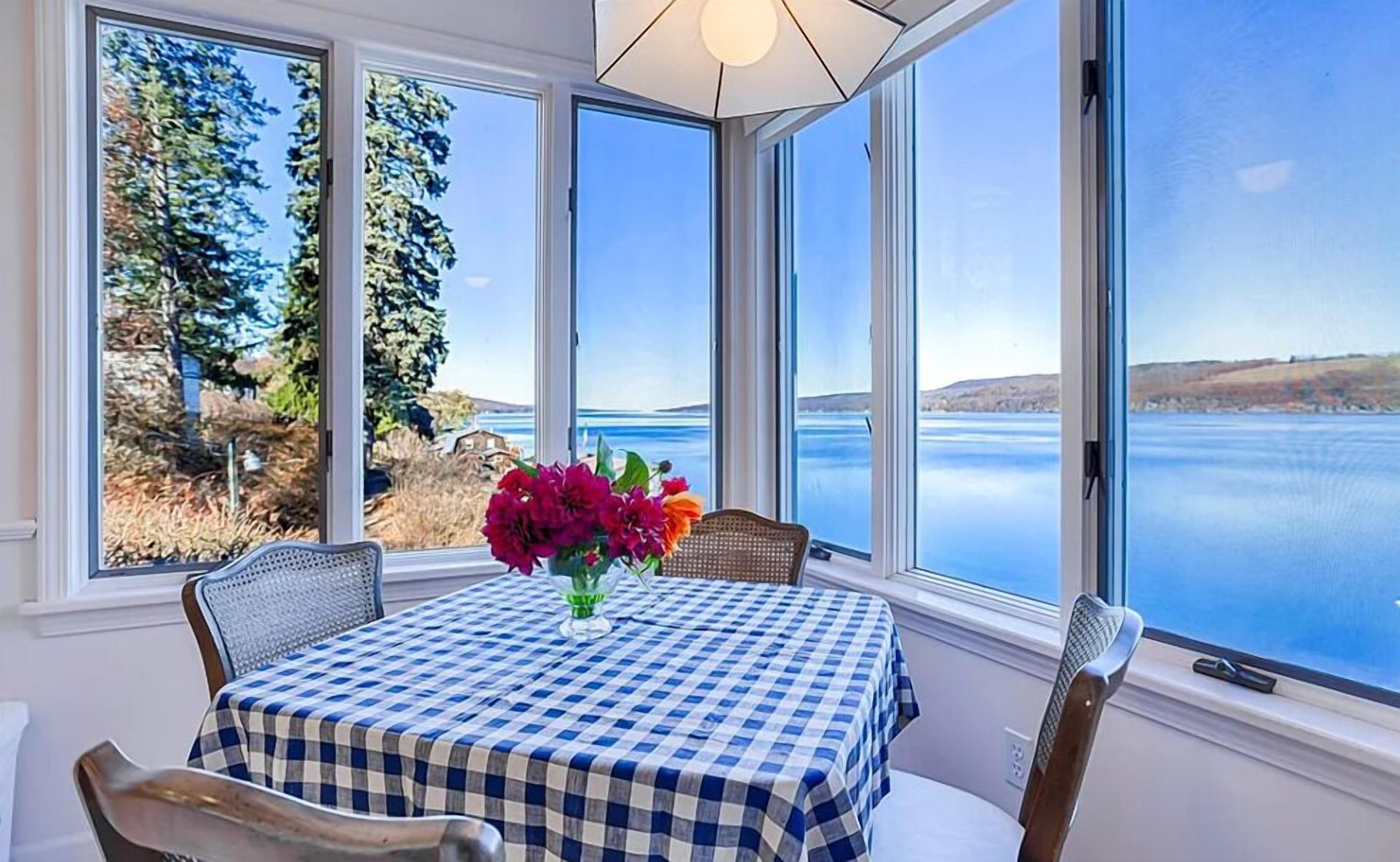 A dining area with a checkered tablecloth, a vase of flowers, and a view of a lake and hills through large windows.