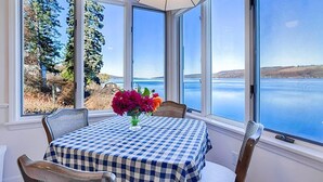 A dining area with a checkered tablecloth, a vase of flowers, and a view of a lake and hills through large windows. - Dean Lake House (Watkins Glen)