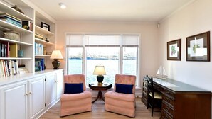 A room with a view of a body of water, a desk with drawers, two chairs, a lamp, and a bookshelf. - Dean Lake House (Watkins Glen)