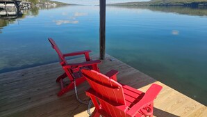 Een houten steiger met twee rode Adirondack-stoelen, een helder uitzicht over een rustig meer en een verre oever met gebouwen en bomen.