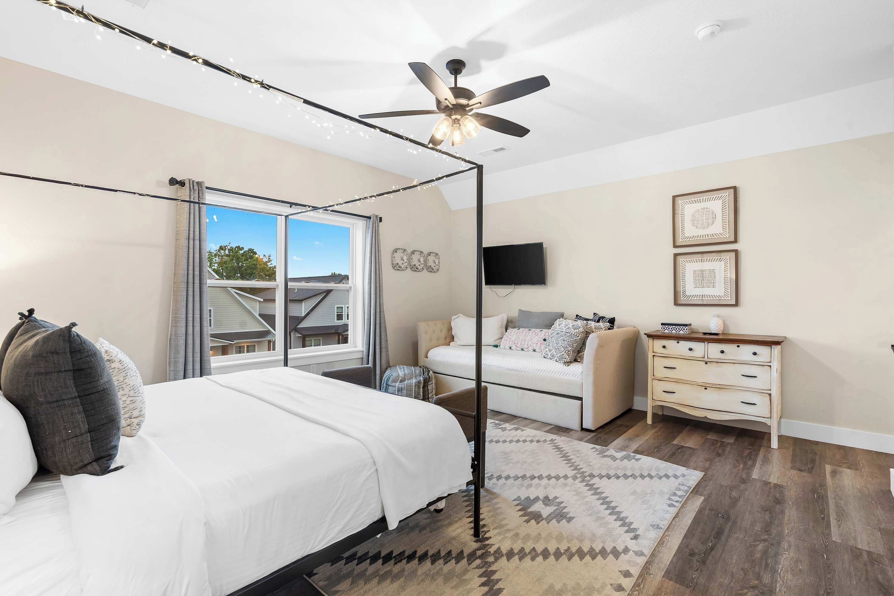 A bedroom with a four-poster bed, a ceiling fan, a large window, a white sofa, a TV, and a dresser.