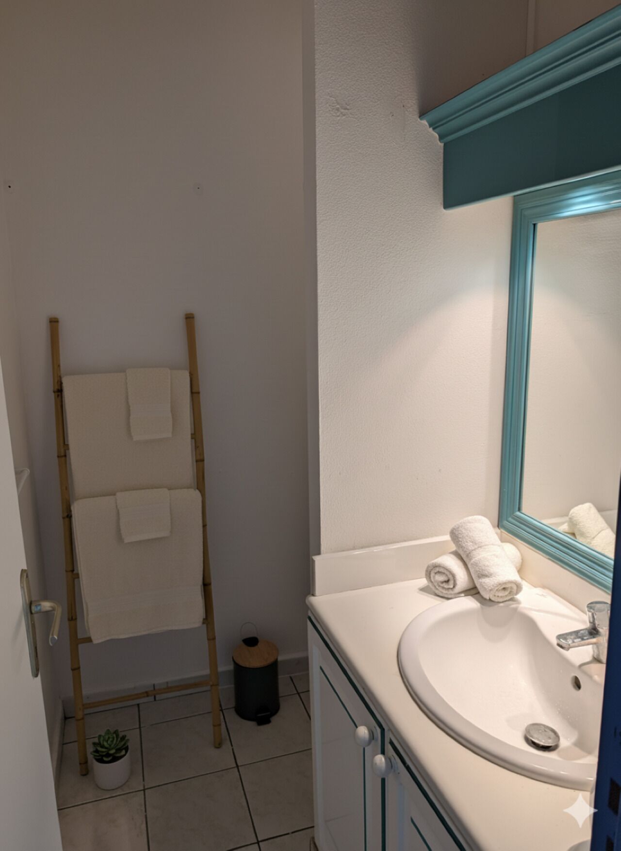 Un baño con lavamanos blanco, un espejo con marco verde azulado y un toallero de bambú.