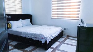 A bedroom with a bed, a bedside table, a window with blinds, and a door. - Terra Santa Lodgings & Restaurants (Adentan)