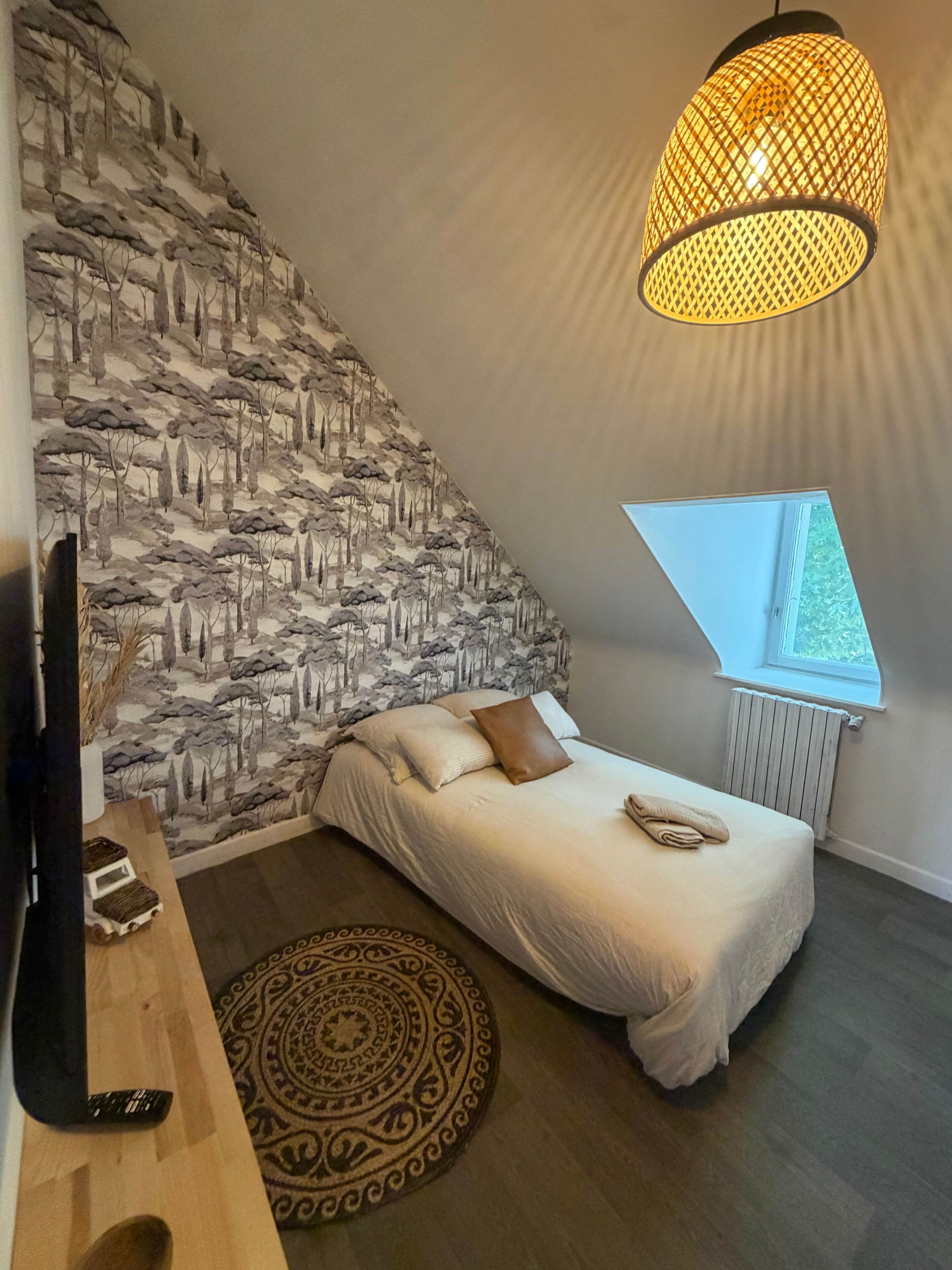 A bedroom with a sloped ceiling, a bed with a white duvet, a patterned wall, a hanging light fixture, and a window with a view.