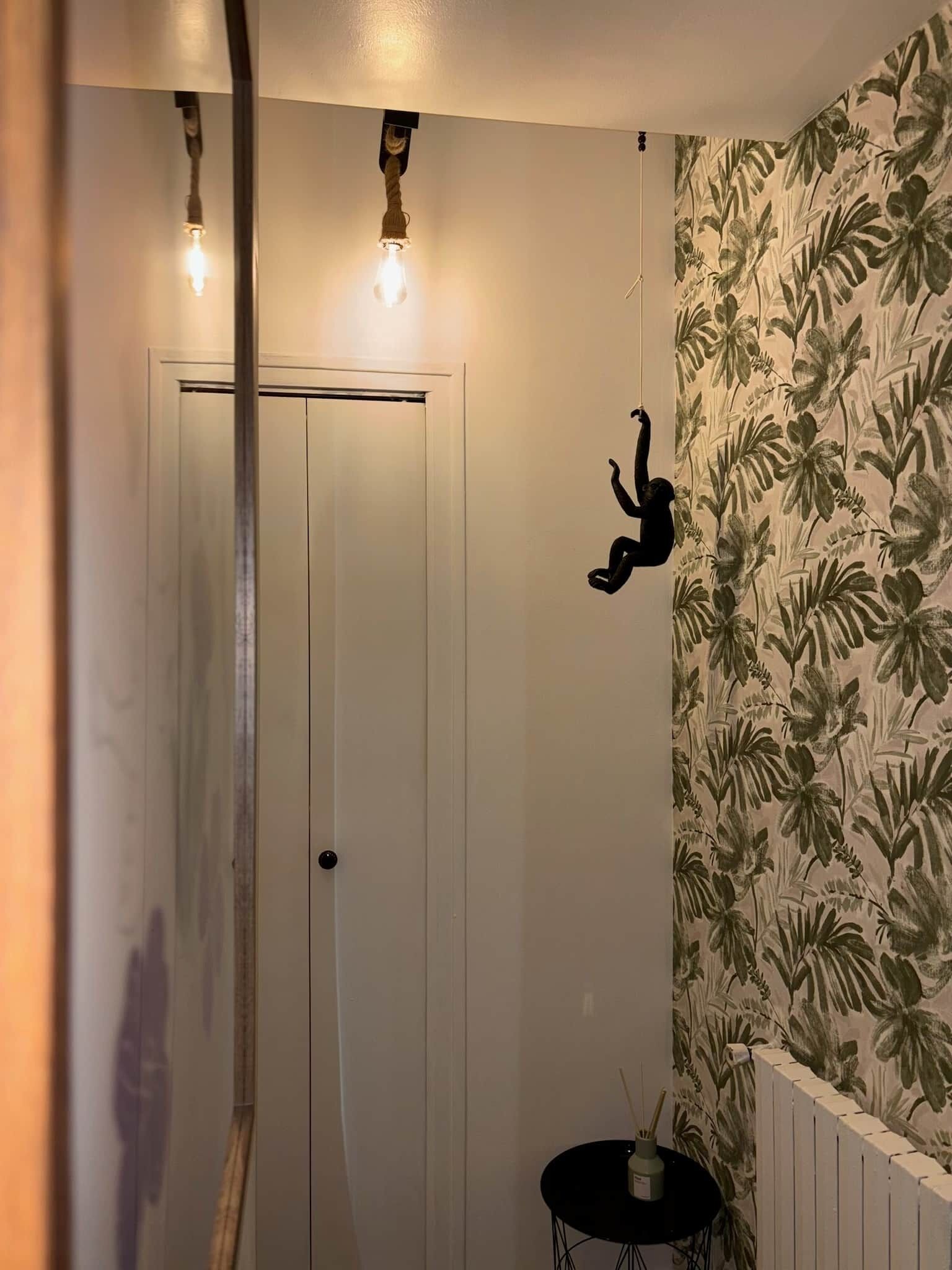 Ein Zimmer mit einer weißen Tür, einer Wand mit tropischem Blattmuster, einem kleinen schwarzen Tisch mit einer Vase und einer Hängeleuchte.