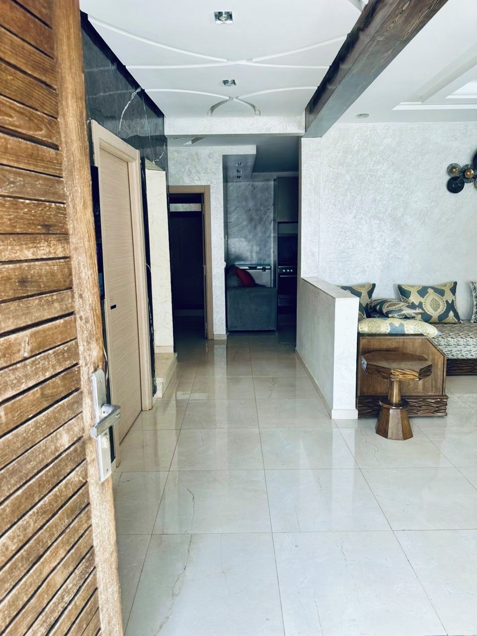 A spacious hallway with a wooden door on the left, a seating area with patterned cushions on the right, and a kitchen in the background.