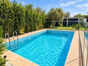 Una piscina rectangular con agua clara y azul, rodeada por un suelo de baldosas y una valla metálica.