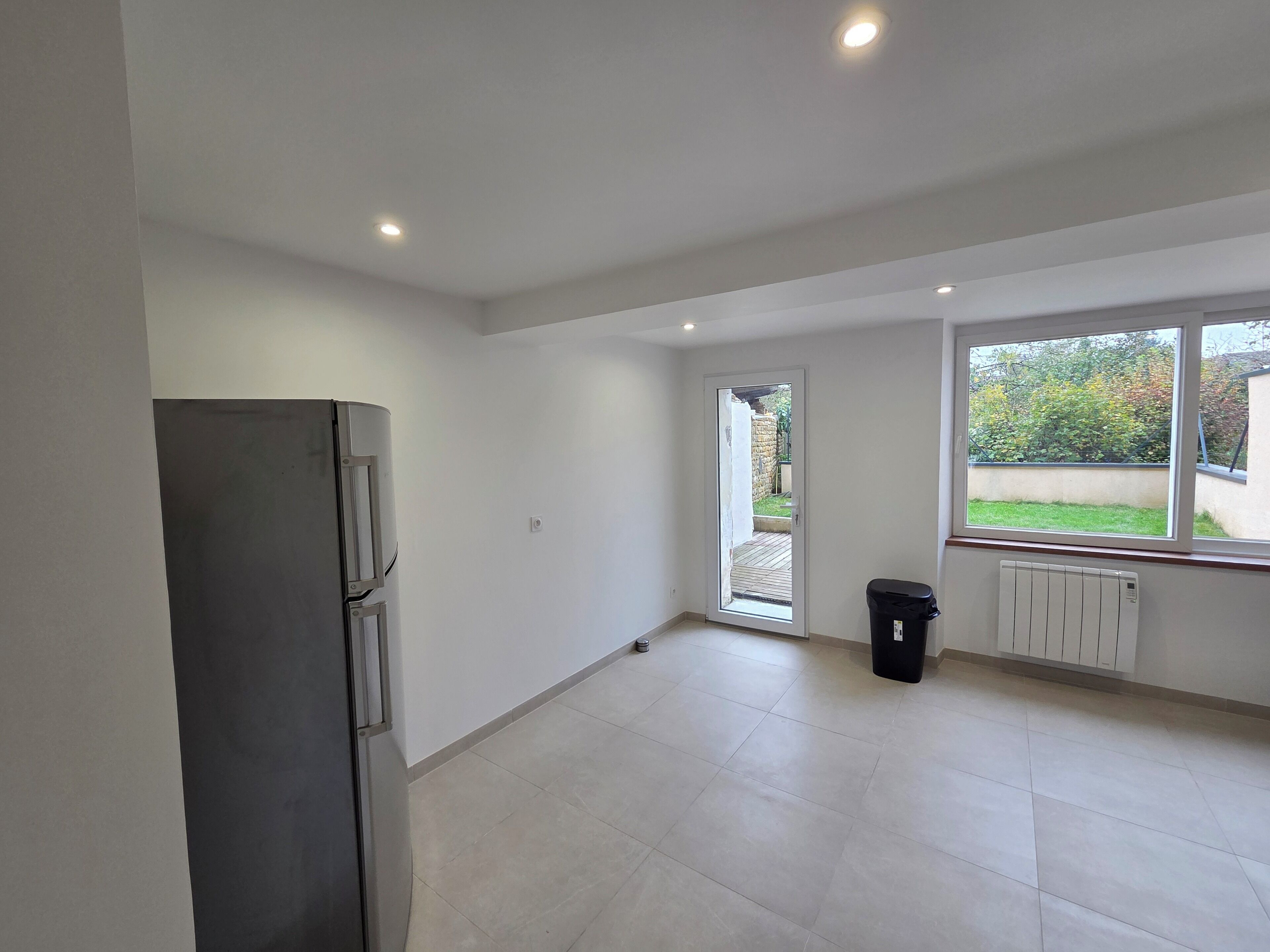 Ruime kamer met een groot raam, een deur naar buiten, een radiator en een prullenbak.