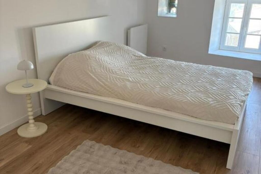 Une chambre avec un lit blanc, une table de chevet ronde et une fenêtre avec des stores.