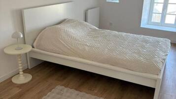 A bedroom with a white bed, a round bedside table, and a window with blinds.