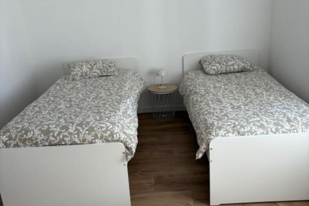 Une chambre avec deux lits simples, chacun agrémenté d’une couvre-lit et de coussins à motifs. On y trouve une petite table ronde avec une lampe, placée entre les deux lits.