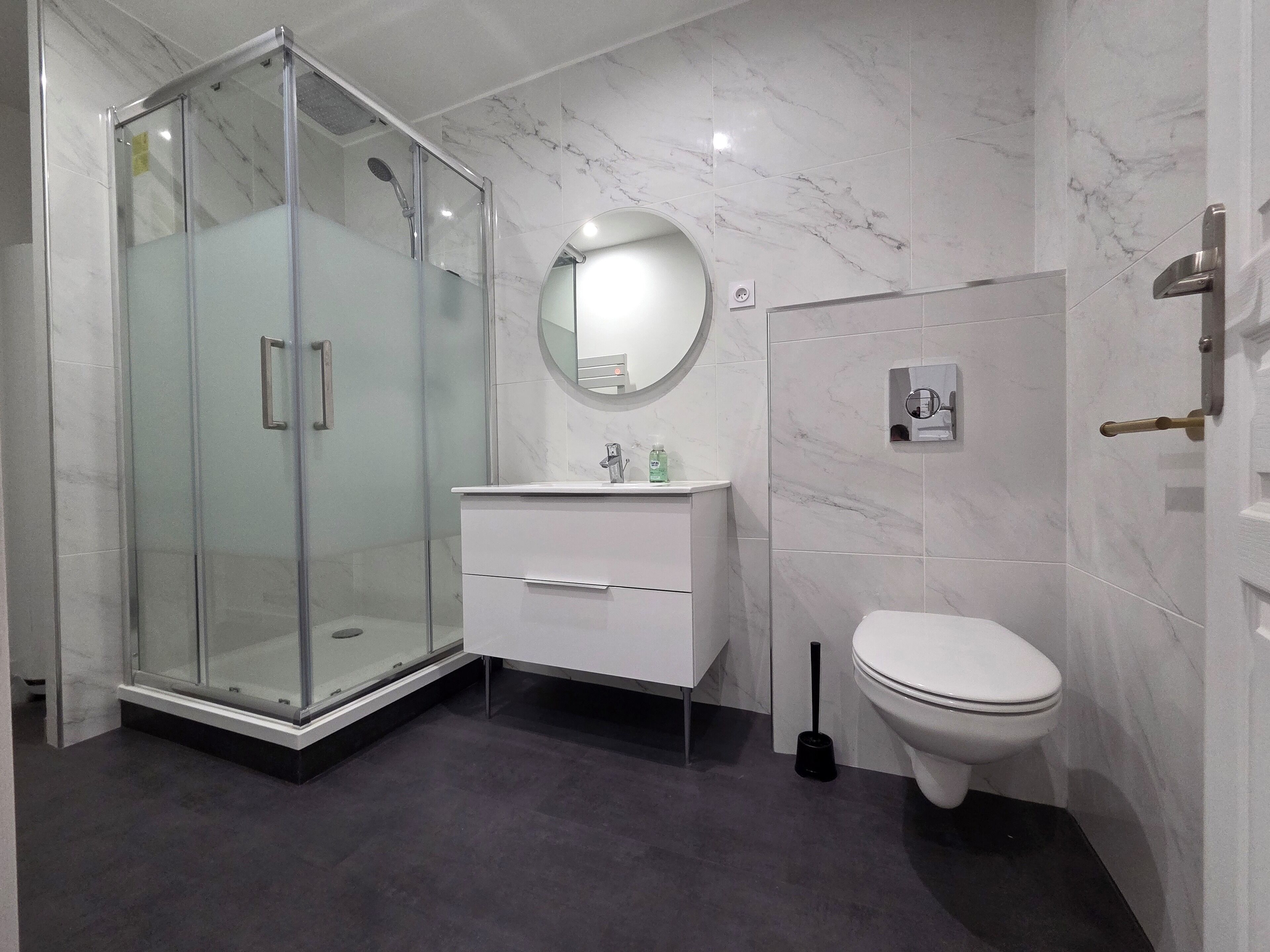 Une salle de bain moderne avec une douche à parois de verre, un meuble-lavabo blanc doté d’un miroir rond et des toilettes.