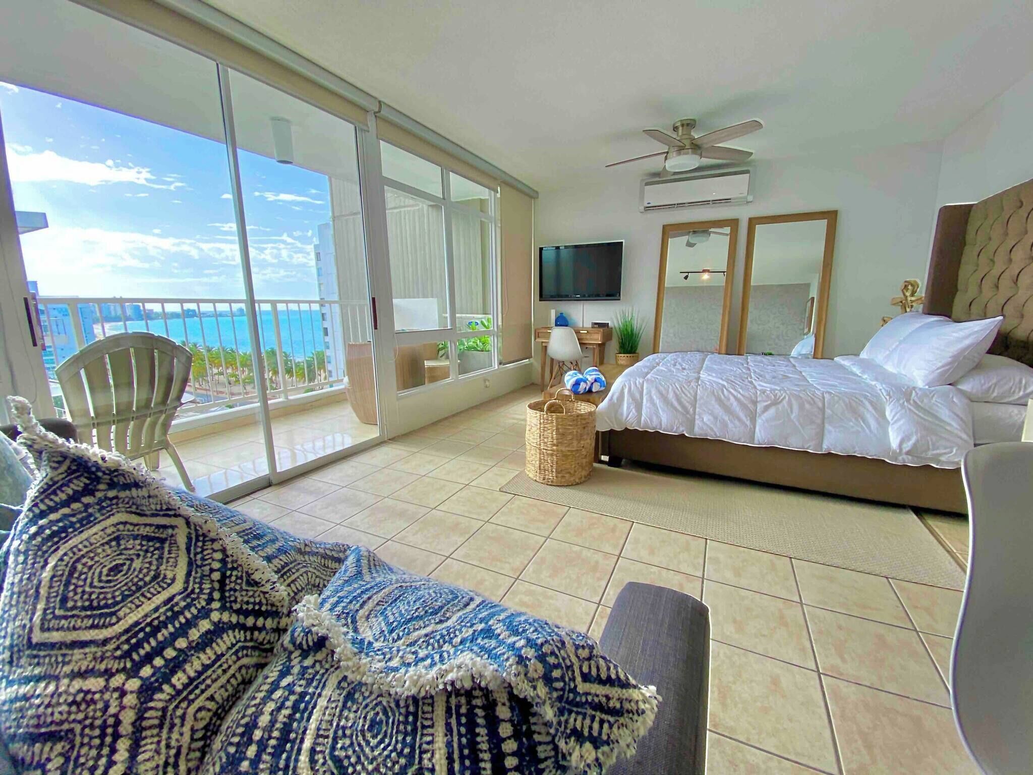 A spacious bedroom with a large bed, a balcony with a view of the ocean, a ceiling fan, and a television.