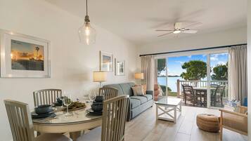 A dining area with a round table, chairs, and a sofa. There are framed pictures on the wall and a ceiling fan above.