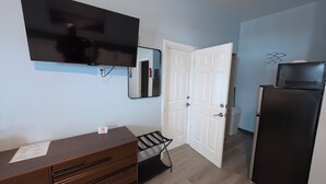 Une pièce avec un téléviseur à écran plat fixé au mur, une porte blanche, un réfrigérateur noir et une commode en bois avec des tiroirs.