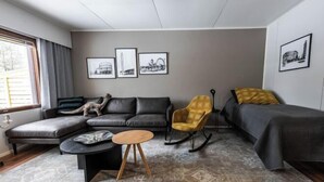 A modern living room with a sectional sofa, a yellow armchair, a small round table, and a bed. There are framed pictures on the wall and a window with curtains. - Cozy Koivutie Apartment (Ranua)