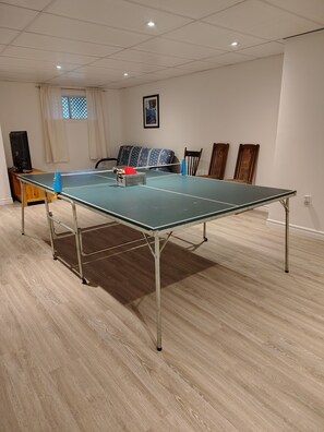 A ping pong table in a room with a wooden floor, a window with curtains, a framed picture on the wall, and a wooden cabinet with a television. - Country house with a forest trail and a mountain bike trail nearby. (Mandeville)
