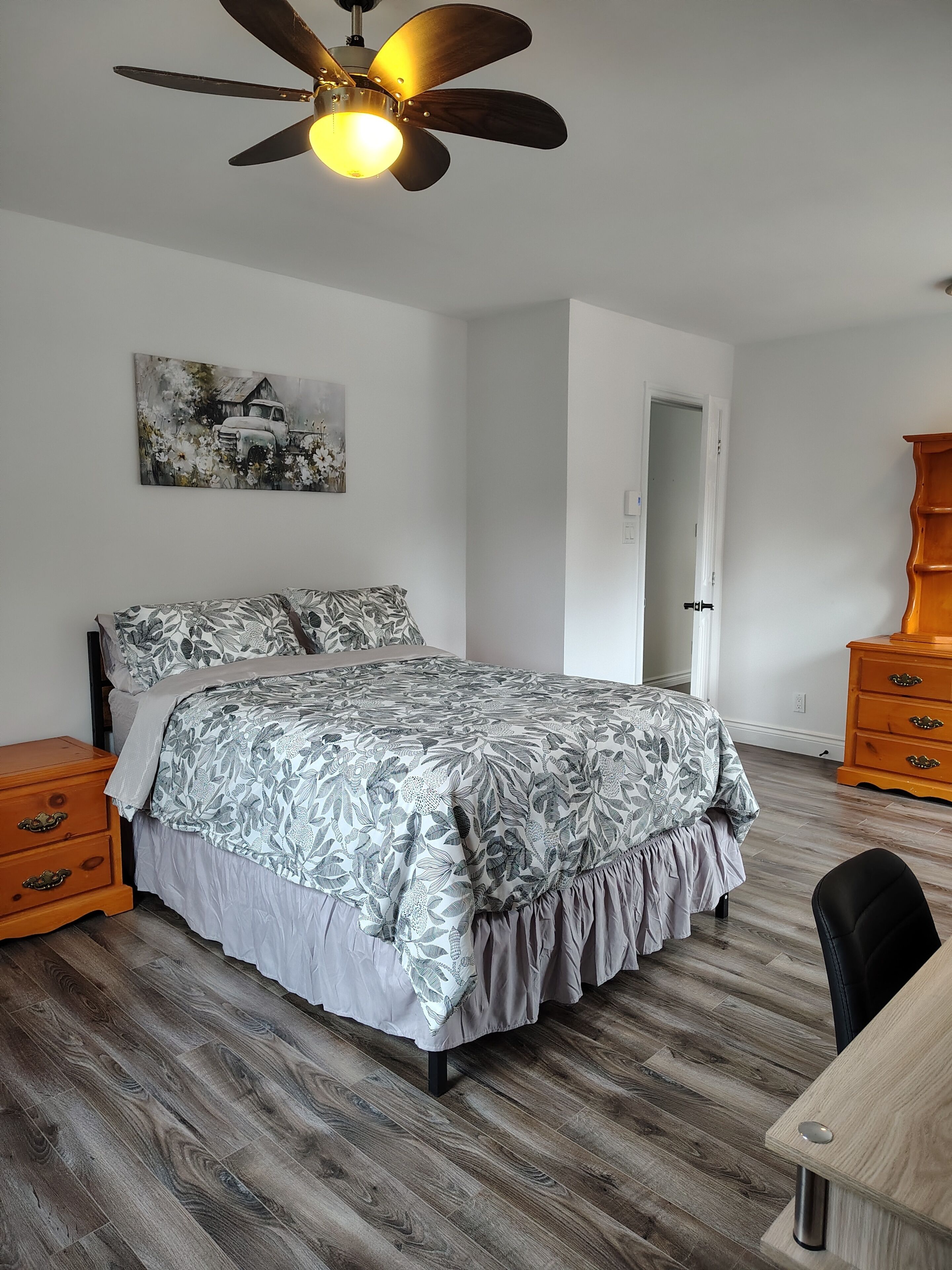 A bedroom with a bed, a ceiling fan, a wooden dresser, and a door.
