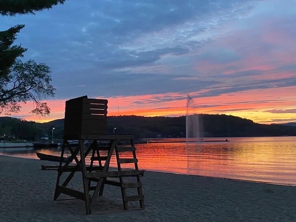 A beach with a wooden chair and a wooden structure, a fountain, and a sunset over water.