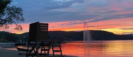 A beach with a wooden chair and a wooden structure, a fountain, and a sunset over water.