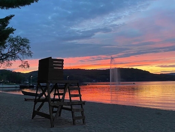 A beach with a wooden chair and a wooden structure, a fountain, and a sunset over water.