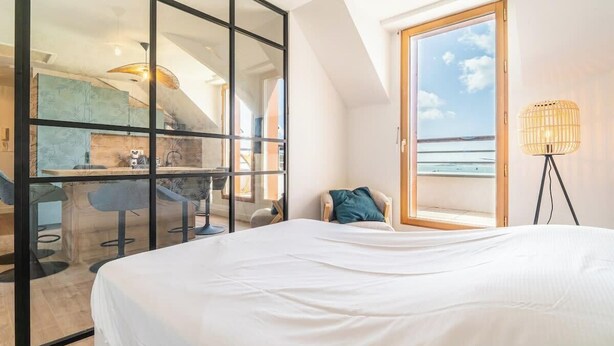 A bedroom with a large bed, a view of the sea through a window, and a kitchenette area visible through a glass partition.