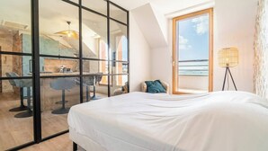 A bedroom with a large bed, a desk, and a chair, a balcony with a view of the sea, and a lamp.