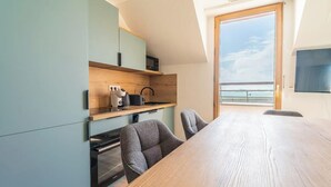 A modern kitchen with a wooden table and chairs, a view of the sea through a large window, and a flat-screen TV mounted on the wall.