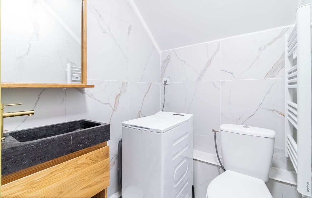 Une salle de bain avec un mur en marbre, un lavabo noir, des toilettes blanches et un lave-linge blanc.