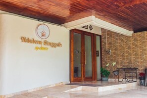 L’entrée d’un hôtel, avec un plafond en bois, une enseigne indiquant « Niklem Singgah » et un mur en briques.