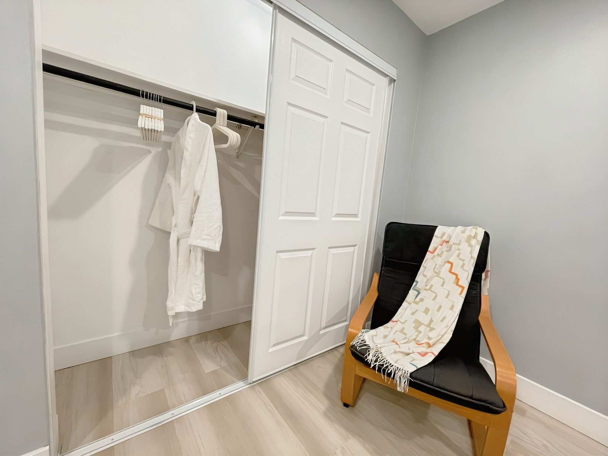 A room with a wooden floor, a white closet with hanging robes, and a black chair with a patterned throw.