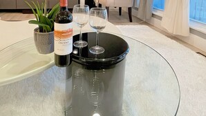 Una sala de estar moderna con una mesa de centro de vidrio, una botella de vino, dos copas de vino, una planta en maceta y un sillón cómodo.